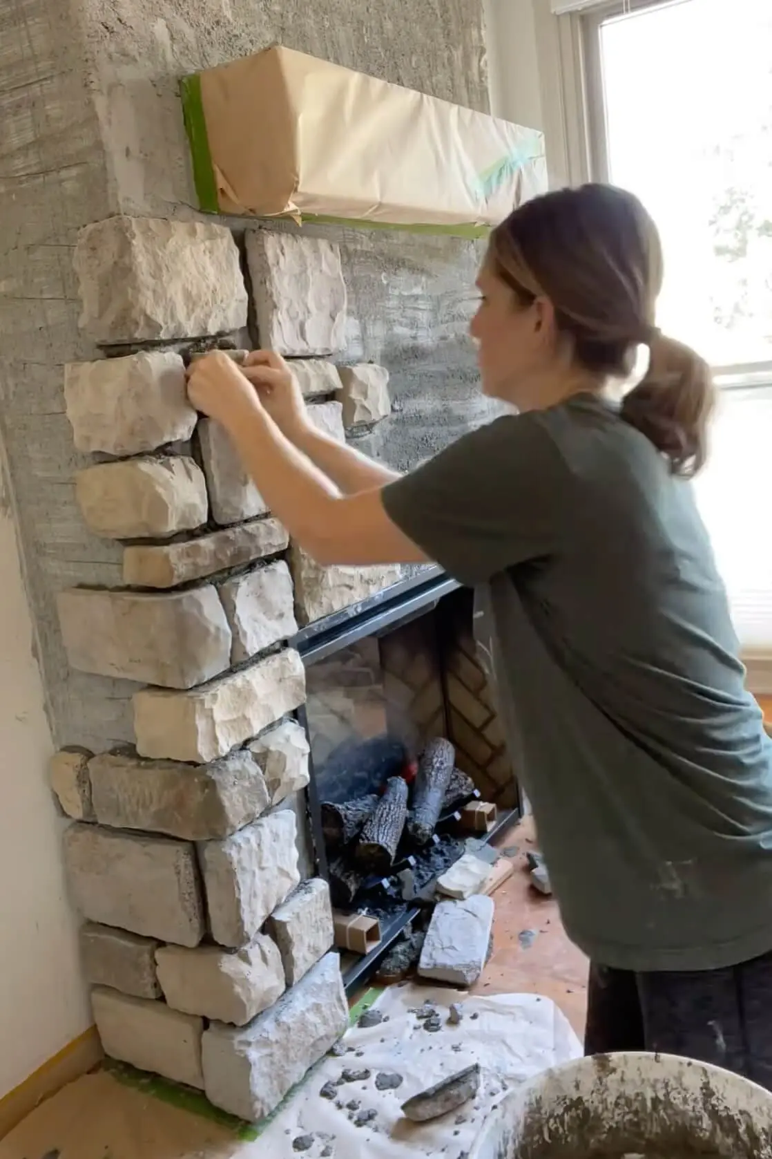Woman carefully placing stacked stone pieces on the side of a fireplace during the DIY installation process.